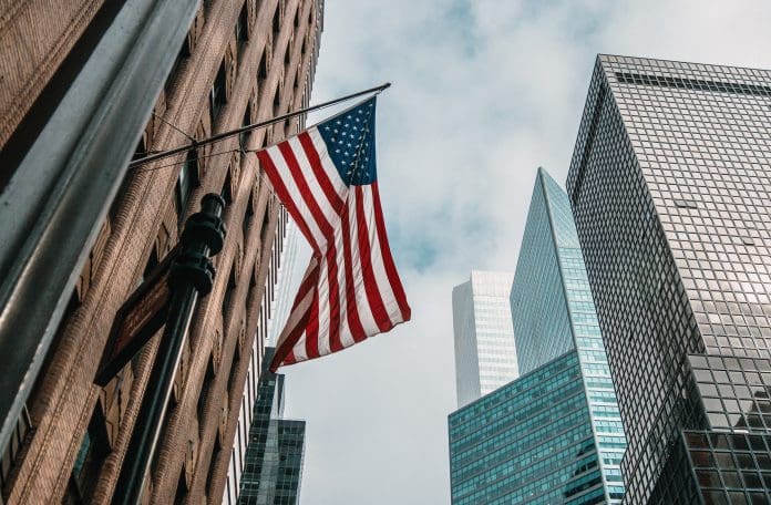 the USA or United States of America flag on a flagpole near skyscrapers under a cloudy sky trump